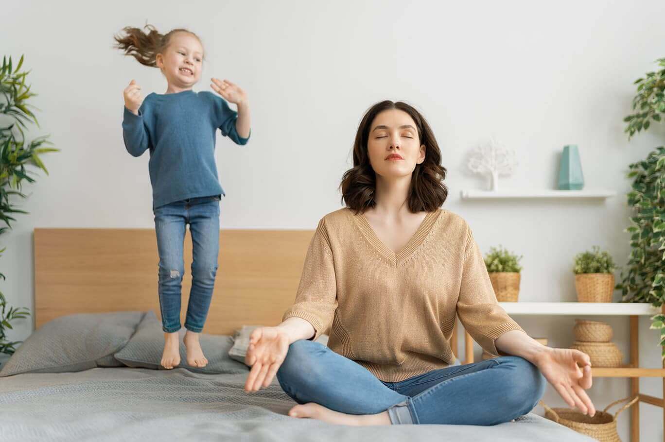 Mutter meditiert auf dem Bett, während im Hintergrund das Mädchen darauf herumspringt.