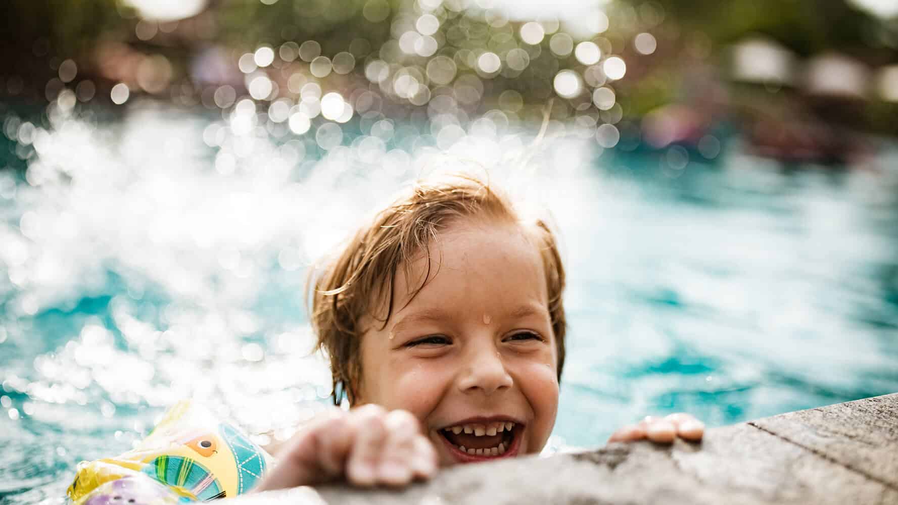 Kind hält sich am Beckenrand und lacht. Es hat Spass im Wasser.