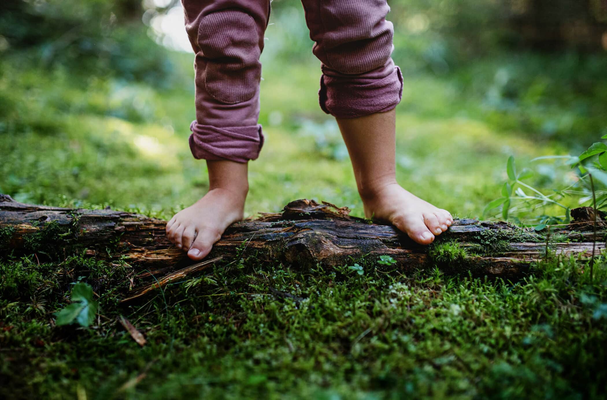 Kind steht barfuss auf einem alten Baumstamm. Es erlebt die Natur mit allen Sinnen.
