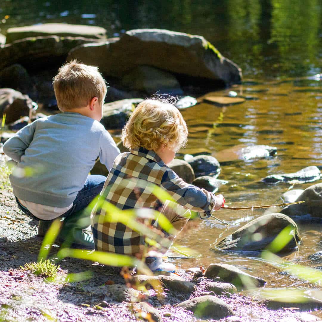 Zwei Jungen spielen am Bach mit Stöcken und dem Wasser. Die Natur ist kostbar und will mit allen Sinnen erlebt werden. Waldtage bei Kimi Krippen fördern dies.